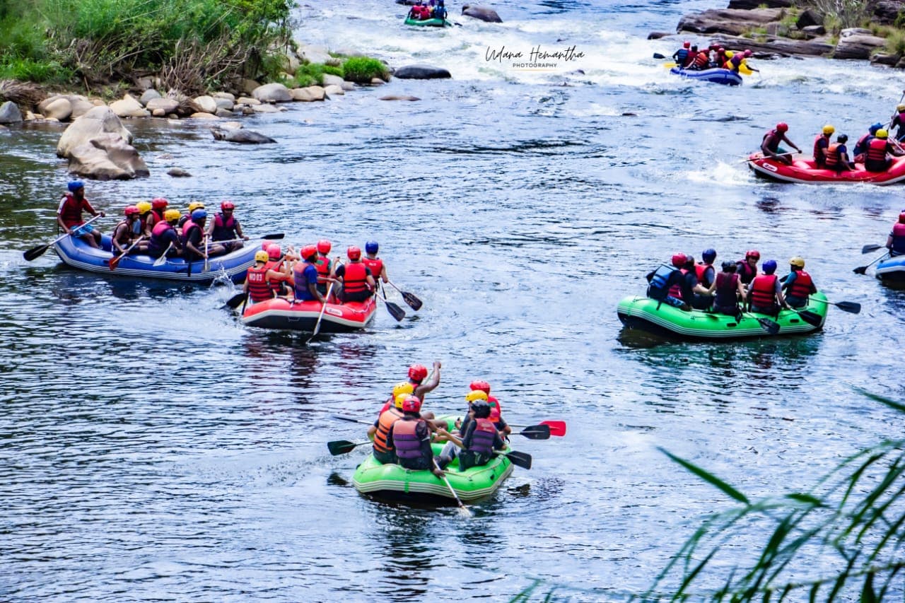Seven rafts carrying a large corporate group down the Kelani River through Kitulgala rainforest, paddles raised in celebration