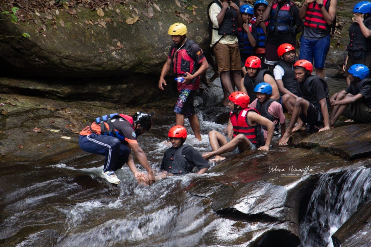 A student group gathered at the base of a waterfall pool on a Kitulgala canyoning route while a guide helps a swimmer