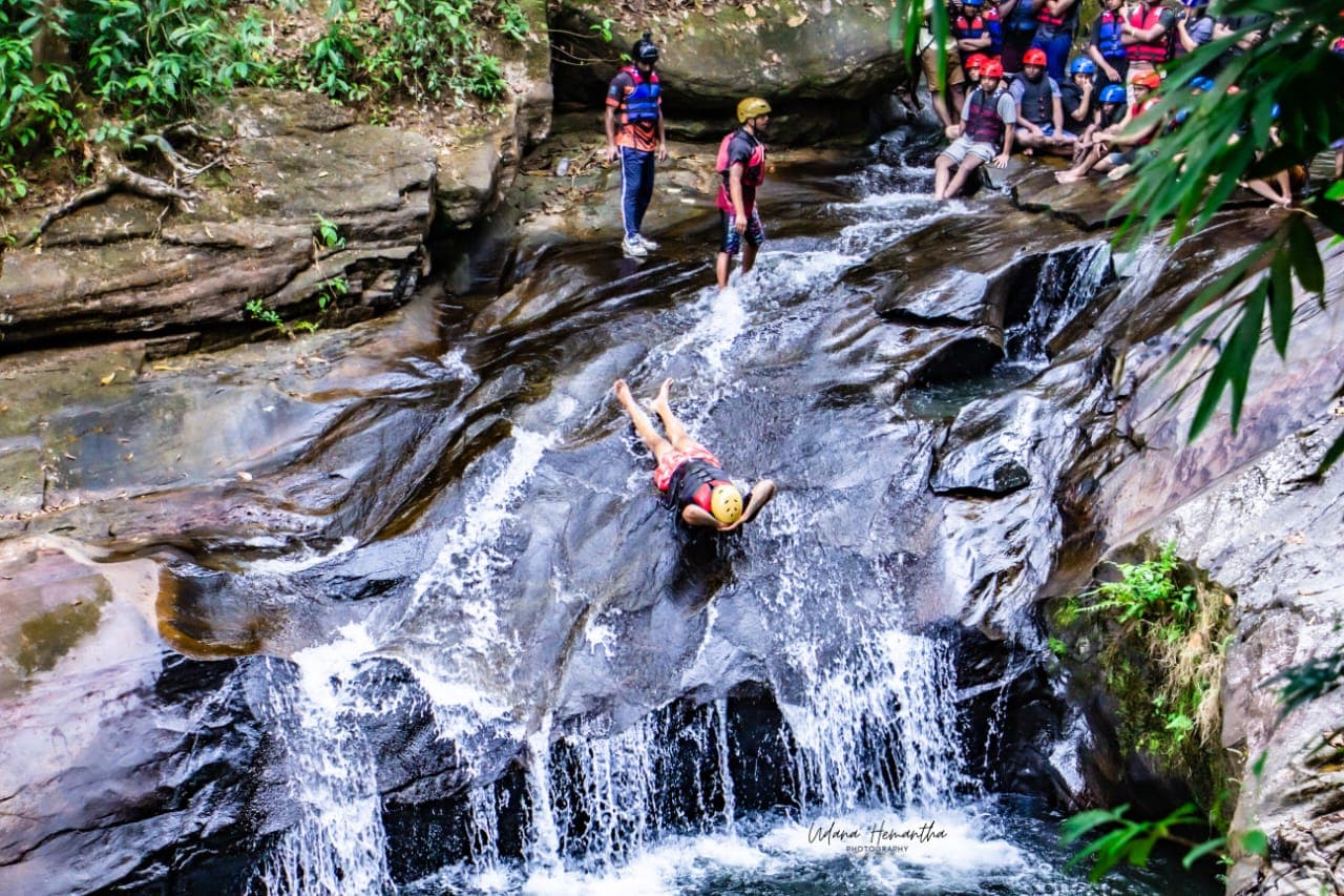 One member of a group riding a natural rock waterslide in the Kitulgala rainforest while the rest of the group watches and cheers