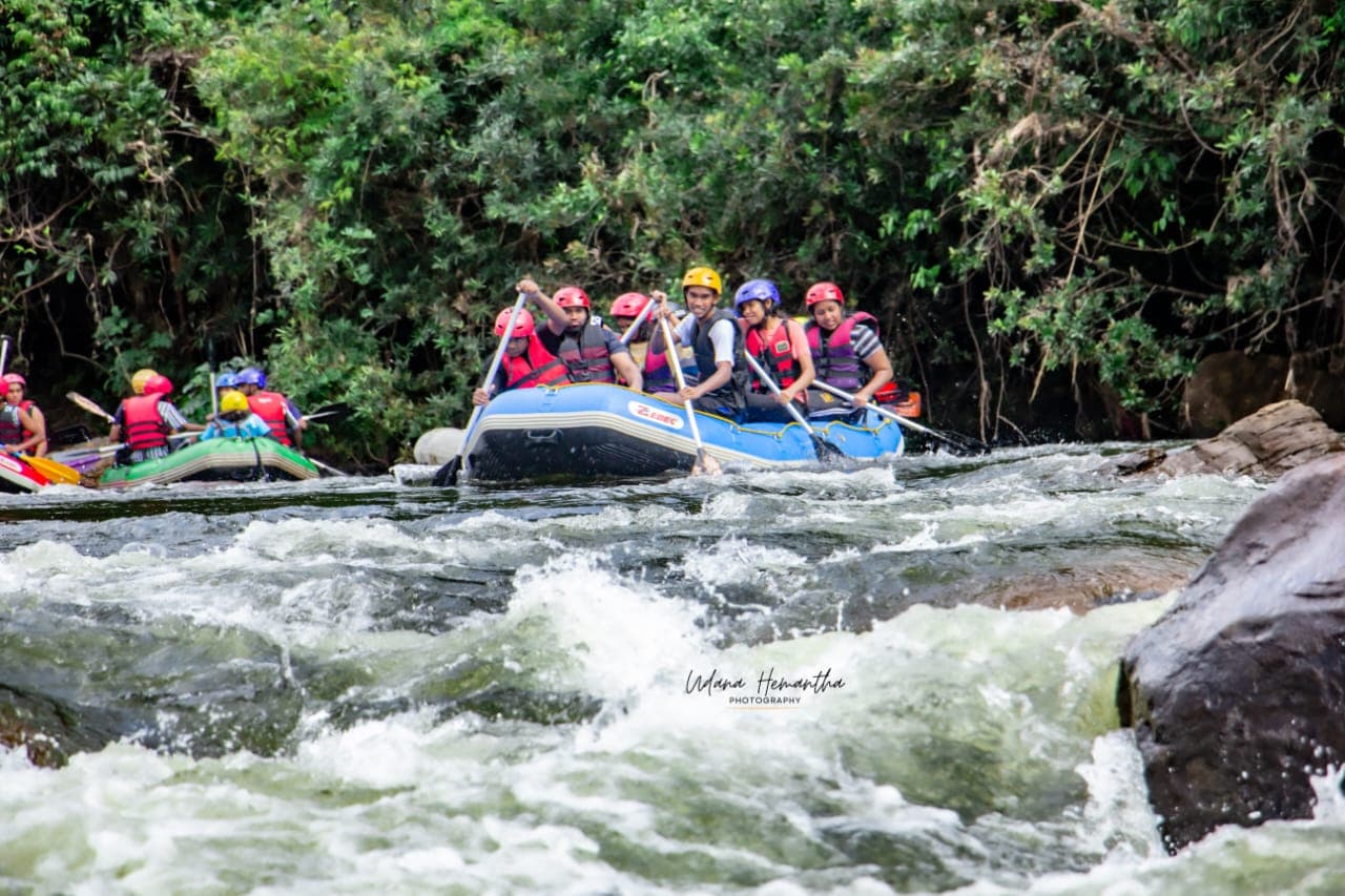 A full raft of teammates paddling towards the camera through white water on the Kelani River, all smiling